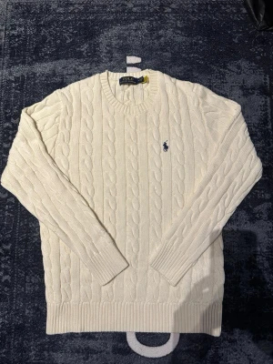 Vit kabelstickad tröja Polo Ralph Lauren - Kabelstickad vit tröja från Polo Ralph Lauren med klassisk blå logga broderad på bröstet. Tröjan har rund halsringning, ribbade muddar och är långärmad. Perfekt för dig som gillar tidlös och clean stil.
