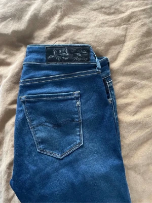 Blå Replay jeans med slim fit - Säljer ett par blå jeans från Replay med klassisk femficksdesign och snygga kontrastsömmar. Jeansen har slim passform och är tillverkade i stretchigt jeansmaterial för extra komfort. Märkespatch bak i midjan och diskreta Replay-detaljer vid fickorna.