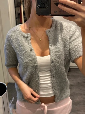 Ljusgrå kofta med pärlknappar - Supermjuk ljusgrå kofta med korta ärmar och pärlknappar framtill. Koftan har en fluffig, stickad känsla och är perfekt att slänga över ett linne. Enkel och stilren design som passar till allt.