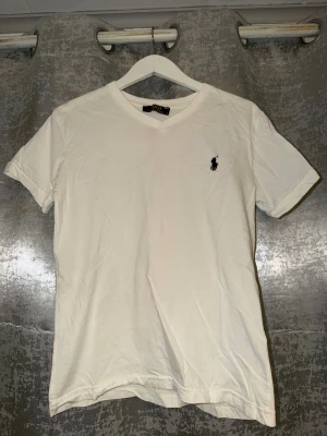 Vit v-ringad t-shirt från Polo Ralph Lauren - Klassisk vit t-shirt från Polo Ralph Lauren med v-ringning och den ikoniska broderade loggan på bröstet. Tillverkad i mjuk bomull och har en normal passform med korta ärmar. Perfekt basplagg för alla tillfällen.