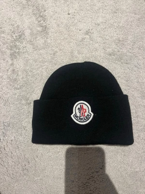 Svart Moncler mössa i ull - Säljer en svart Moncler mössa med klassisk uppvikt kant och broderad logotyp framtill. Mössan är ribbstickad och gjord i mjuk ull som håller dig varm under kalla dagar. Perfekt accessoar för dig som vill ha en clean och stilren look.