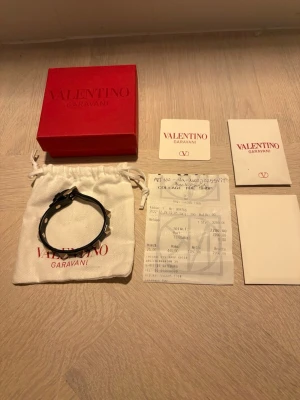 Valentino Garavani Rockstud-armband - Elegant armband i svart läder med Rockstud-nitar i guldfärgad metall. Justerbar storlek och tidlös design som passar både till vardag och fest. Levereras med originalbox, dustbag, kort och kvitto.