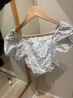 Blommig offshoulder topp från H&M - Supersöt offshoulder topp från H&M med puffärmar och croppad passform. Vit botten med små blå blommor över hela tyget. Tillverkad i ett lätt och luftigt material, perfekt för varma dagar. Passar dig som gillar romantisk och trendig stil.