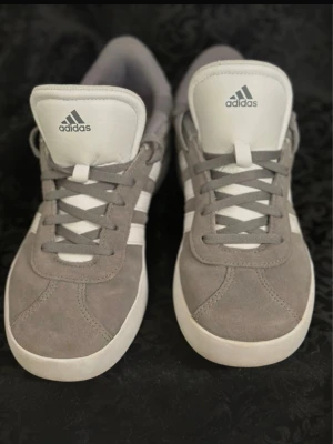 Gråa Adidas sneakers i mocka 38 - Säljer ett par stilrena Adidas sneakers i grå mocka med vita detaljer och klassiska tre ränder på sidan. Skorna har snörning och vit sula, samt Adidas-logga på plösen. Perfekta för dig som gillar en clean och sportig look.