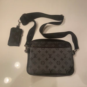 Louis Vuitton axelväska monogram svart - Säljer en snygg axelväska från Louis Vuitton i svart med klassiskt monogrammönster. Väskan har justerbar axelrem, flera fack med dragkedja och en liten avtagbar plånbok. Tillverkad i tåligt skinn med silverfärgade detaljer.