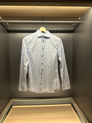 Ljusblå randig skjorta Riley Slim Fit - Säljer en ljusblå skjorta från Riley i slim fit-modell. Skjortan har tunna vita ränder, klassisk krage och svarta knappar hela vägen fram. Tillverkad i bomull och har långa ärmar med knapp vid manschetten. Perfekt för dig som gillar stilrena och fräscha plagg.