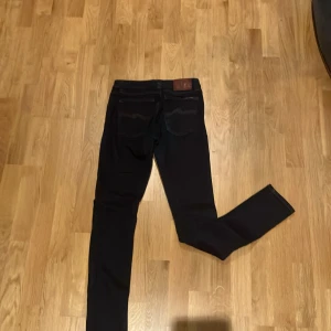 Svarta jeans från Nudie Jeans, 26/32 - Säljer ett par svarta jeans från Nudie Jeans i storlek 26/32. Klassisk femficksmodell med snygga detaljer på bakfickorna och läderpatch i midjan. Jeansen har smal passform och är tillverkade i slitstarkt denimtyg. Modell: Sam