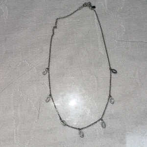 Äkta Silverhalsband med glittriga hängen - Ett tunt silverfärgat halsband med små ovala hängen dekorerade med glittrande stenar. Kedjan är smal och smidig, och hänget har en droppformad detalj vid låset. Perfekt för dig som gillar stilrena och glittriga accessoarer.