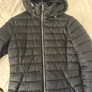 Svart pufferjacka från Zara med huva - Svart pufferjacka från Zara med horisontella sömmar, dragkedja framtill och praktisk huva med dragsko. Jackan har två snedställda fickor och är tillverkad i ett lätt, syntetiskt material som håller värmen under kyliga dagar.