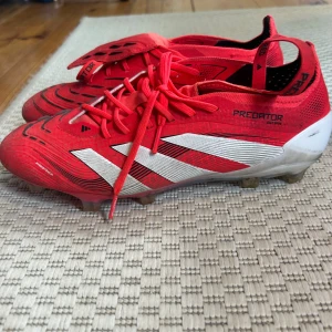 Adidas Predator elite röda fotbollsskor - Säljer ett par röda Adidas Predator elite fotbollsskor med vita och svarta detaljer. Skorna har snörning, låg profil och mönstrad ovandel för extra grepp. Snygg design med klassiska tre ränder på sidan och vit häl. Perfekta för dig som vill dominera på planen. Använt dem i 3 månader och nytt pris är 3400 kr. 