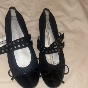 Svarta ballerinaskor  - Snygga svarta ballerinaskor från Cider med lackad tå, dekorativa rosetter och dubbla remmar med nitar och spännen i metall. Skorna har rund tå och platt sula, vilket ger en klassisk men edgy look. Perfekta för att lyfta vilken outfit som helst.