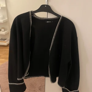 Svart kofta från Gina Tricot - Svart kofta från Gina Tricot med kontrasterande vita sömmar längs kanterna. Koftan har en öppen front och långa ärmar, perfekt för lager-på-lager. Enkel och stilren design som passar till det mesta.