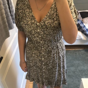 Svartvit blommig kortklänning med knappar - Superfin svartvit kortklänning från H&M med blommigt mönster, v-ringning och knappar framtill. Klänningen har korta ärmar och volangkant nertill. Perfekt för varma dagar och enkel att styla med sneakers eller sandaler.