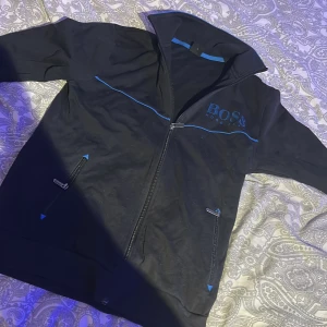 Svart jacka från Hugo Boss med blå detaljer - Svart jacka från Hugo Boss med blå detaljer och logga på bröstet. Jackan har hel dragkedja, ståkrage och två fickor med dragkedja. Snygg och sportig look med blå piping längs bröstet och insidan av kragen.