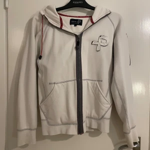 Vit hoodie från Pelle P med dragkedja - Vit hoodie från Pelle P med grå kontrastsömmar, hel dragkedja och huva med röd snörning. Stort P-logga på bröstet och broderad detalj på ärmen. Praktiska fickor framtill och sportig känsla. Perfekt för chill eller utomhusaktiviteter.