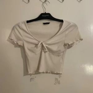 Vit croppad ribbad topp med knyt - Säljer en vit croppad topp från KappAhl med ribbad struktur och söt knytning framtill. Toppen har korta ärmar med vågig kant och djup v-ringning. Perfekt till jeans eller kjol för en chill och trendig look.