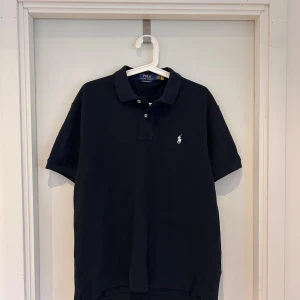 Mörkblå pikétröja från Ralph Lauren - Snygg mörkblå pikétröja från Ralph Lauren med klassisk krage, korta ärmar och vit broderad logga på bröstet. Modellen är custom slim fit och tillverkad i mjuk bomull. Perfekt för en clean och stilren look.