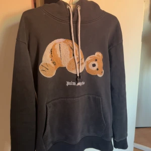 Svart hoodie Palm Angels med nalle - Svart hoodie från Palm Angels med stor broderad nalle på bröstet och vit logga under. Tröjan har huva med snörning och en stor magficka. Perfekt för dig som gillar streetwear och vill sticka ut med en unik detalj.