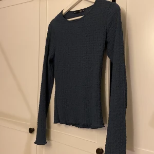Mörkblå långärmad topp från lager 157 - Mörkblå långärmad topp från lager 157 i storlek XS. Toppen har en strukturerad, ribbad yta och rund halsringning. Modellen är figurnära och har lätt vågiga kanter vid ärmslut och nederkant. Perfekt för dig som gillar enkel men snygg stil. Helt oanvänd 