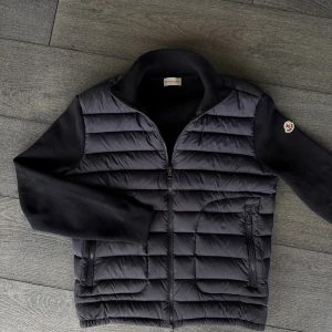 Marinblå Moncler cardigan  - Svart pufferjacka från Moncler med quiltad front och stickade ärmar. Jackan har dragkedja framtill, två fickor med dragkedja och Moncler-logga på ena ärmen. Snygg och stilren design som passar perfekt till höst och vinter.