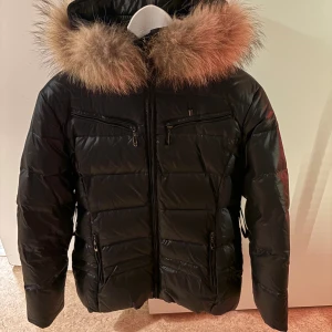 Svart pufferjacka med fuskpäls från Alvaro - Säljer en svart pufferjacka från Alvaro med fluffig fuskpäls runt huvan och leopardmönstrat innerfoder. Jackan har dragkedjor framtill och flera fickor, perfekt för kalla dagar. Snygg och trendig look med coola detaljer.