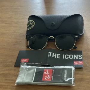 Svarta Ray-Ban Clubmaster solglasögon - Ray-Ban Clubmaster solglasögon med svart båge och guldiga detaljer. Klassisk retroform med halvbåge och mörka linser. Levereras med originalfodral, putsduk och manual. Perfekt för dig som vill ha en ikonisk och tidlös look.