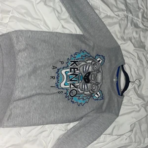 Grå Kenzo sweatshirt med tigertryck - Säljer en grå sweatshirt från Kenzo med ikoniskt tigerbroderi i blått, svart och vitt framtill. Tröjan har rund hals, långa ärmar och är tillverkad i mjuk bomull. Perfekt statement-piece för dig som gillar streetstyle och vill sticka ut.