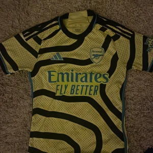 Arsenal x Adidas gul bortatröja XS - Säljer en Arsenal bortatröja från Adidas i storlek XS. Tröjan är gul med svarta vågiga ränder och små svarta prickar, samt blå detaljer och broderat klubbmärke. Tillverkad i lätt och ventilerande polyester, perfekt för match eller träning.