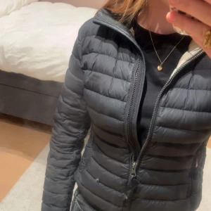 Svart dunjacka Parajumpers YS - Snygg svart dunjacka från Parajumpers i storlek YS. Jackan är quiltad med dragkedja framtill, två sidofickor med dragkedja och klassisk logga på ärmen. Insidan är ljus och jackan har en smal passform som sitter tajt och stilrent. Jackan har även ett litet hål längst ner men det skulle kunna gå att laga för någon med kunnighet.
