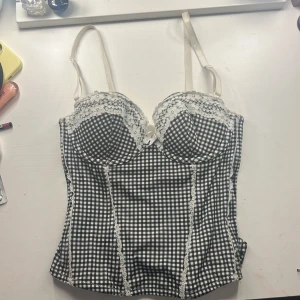 Rutig svartvit korsett med spets - Supersöt korsett med svartvitt rutigt mönster och vita spetsdetaljer vid kuporna och längs sömmarna. Justerbara axelband och hakknäppning bak. 