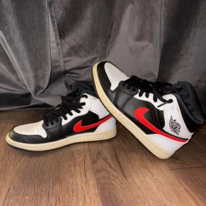 Nike Air Jordan 1 Chile Red Mid. St.40 - Nike Air Jordan 1 sneakers i svart, vitt och rött med klassisk röd swoosh på sidan. Skorna har snörning, rund tå och platt sula. Tillverkade i skinn med coola kontraster och ikonisk Jordan-logga på hälen. Perfekt för dig som gillar streetstyle och vill sticka ut.