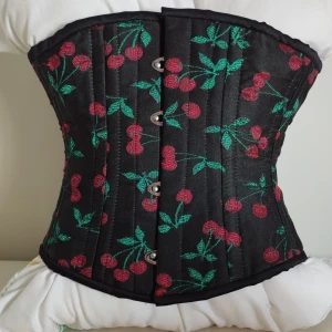 Underbystkorsett double steelboning körsbär - Snygg svart korsett med broderade röda körsbär. Klassik knäppning fram och snörning baktill. Korsett storleken är 20 vilket betyder att du kan snöra ned din midja till 50cm och få en dramatisk timglasmidja. Materialet är glansigt och känns lyxigt, perfekt för daglig användning i Lolita eller retro stil, eller för en fest. Den är från Orchard corsettes för 1000kr, men jag tog bort lappen. Den har burits en gång utanpå kläder.