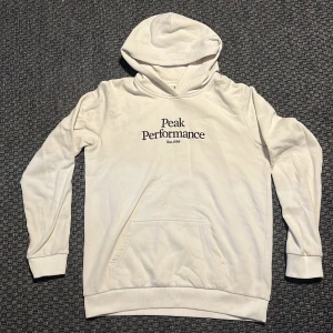 Vit hoodie från Peak Performance - Säljer en stilren vit hoodie från Peak Performance med svart broderad logga på bröstet. Tröjan har huva, magficka och långa ärmar. Perfekt för dig som gillar en clean och klassisk look.