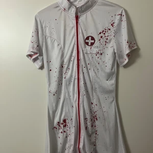 Vit sjuksköterskekläning med blodstänk - Vit kortklänning i 100% polyester med röda blodstänk och ett sjukvårdsemblem på bröstet. Klänningen har korta ärmar, röd dragkedja framtill och röda detaljer längs sömmarna. Perfekt för temafest eller halloween!