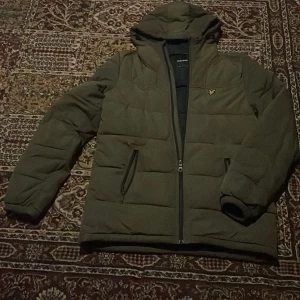 Olivgrön pufferjacka från Lyle & Scott - Säljer en olivgrön pufferjacka från Lyle & Scott med huva och dragkedja framtill. Jackan har två fickor med dragkedja och den klassiska Lyle & Scott-loggan på bröstet. Perfekt för kyliga dagar och riktigt skön att bära.