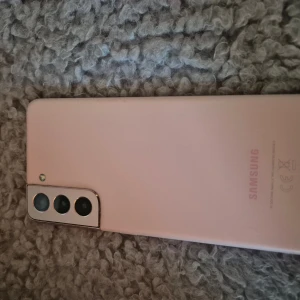 Samsung Galaxy S21 5g - Samsung Galaxy S21 5g i rosa färg med trippelkamera. Skärmen ser ut att vara i gott skick utan synliga sprickor. Mindre slitage syns på ramen, men inget som påverkar funktion. Perfekt för dig som vill ha en kraftfull Android-telefon med snygg design.