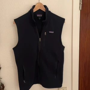 Mörkblå fleeceväst från Patagonia - Mörkblå fleeceväst från Patagonia med hög krage och hel dragkedja framtill. Västen har två fickor med dragkedja och en liten logotyp på bröstet. Perfekt för lager-på-lager och outdoor-vibes.
