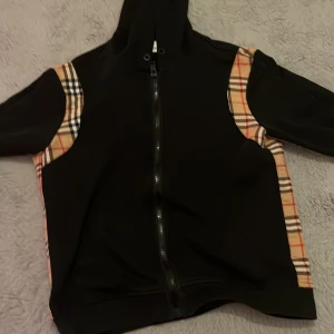 Svart hoodie med rutiga detaljer - Svart hoodie från Burberry med klassiska beige, röda och svarta rutiga paneler längs ärmar och sidor. Tröjan har dragkedja framtill, huva och långa ärmar. Perfekt för dig som vill ha en stilren men ändå unik look.