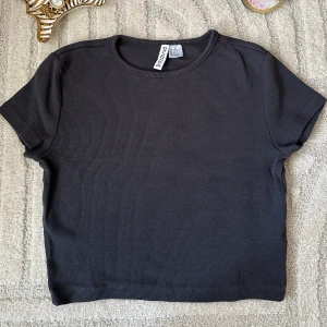 Mörkgrå ribbad topp från Monki - En stilren mörkgrå ribbad topp från Monki med korta ärmar och rund halsringning. Perfekt att matcha med jeans eller kjol för en enkel och snygg look. Materialet är mjukt och stretchigt för extra komfort.