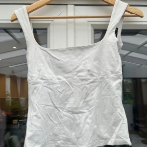 Vit linnetopp med knyt i ryggen - Säljer en stilren vit linnetopp med breda axelband och fyrkantig ringning. Toppen har en snygg knytning i ryggen som ger en extra detalj. Perfekt till jeans eller kjol för en fräsch look. Tillverkad i mjuk bomull som känns skön mot huden.