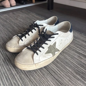 Golden Goose Superstar sneakers  - Golden Goose Superstar sneakers i vitt skinn med svarta snören och mörkblå hälkappa. Sidan med stjärnan i olivgrönt. 42 passar 43. Skriv till mig för förhandling av pris. 