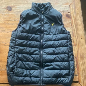 Svart dunväst från Lyle & Scott Junior - Svart quiltad dunväst från Lyle & Scott Junior med gul logga på bröstet. Västen har hög krage, dragkedja framtill och två fickor. Perfekt för lager-på-lager och enkel att matcha med hoodies eller tröjor.