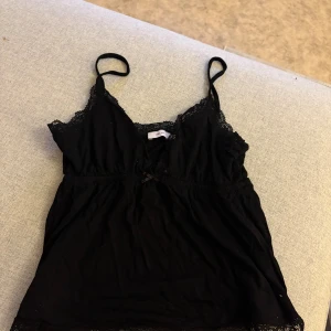 Svart spetslinnetopp från Nelly - Svart linne från Nelly med tunna axelband och v-ringning. Toppen har fina spetsdetaljer längs urringningen och nederkanten samt en liten rosett framtill. Mjuk och lätt bomullskänsla, perfekt för en chill och snygg look.