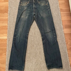 Levi's 501 blå jeans straight fit - Klassiska blå Levi's 501 jeans med raka ben och femficksmodell. Jeansen har en snygg tvätt med slitningar och den ikoniska läderpatchen bak i midjan. Perfekta för en avslappnad och tidlös look.