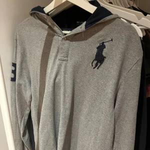 Grå hoodie från Polo Ralph Lauren - Grå långärmad hoodie från Polo Ralph Lauren med marinblå huva och stor broderad logga på bröstet. Pikéknäppning framtill och siffra på ärmen. Skön bomullskvalitet, perfekt för chill dagar.                                                    Barnstorlek XL men sitter som vanlig S