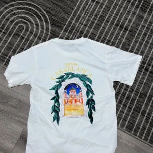 Vit t-shirt från Casablanca med tryck  - Vit t-shirt från Casablanca med färgglatt tryck av en byggnad och gröna blad både fram och bak. Klassisk rund hals och korta ärmar. Perfekt för dig som gillar snygga prints och vill sticka ut lite extra. Gjord i mjuk bomull för skön känsla hela dagen.  Den passar M också 