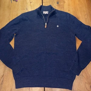 Marinblå stickad tröja från Morris - Snygg marinblå stickad tröja från Morris med half zip och ribbade muddar. Tröjan har en liten vit broderad logga på bröstet och hög krage. Perfekt för dig som gillar klassisk och stilren look.