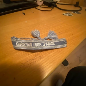 Blått Christian Dior J'Adior armband - Snyggt blått armband från Christian Dior med texten 'J'Adior' broderad i vitt. Armbandet har justerbar knytning med två tofsar i ändarna och är gjort i ett mjukt tygmaterial. Perfekt accessoar för att lyfta din outfit med en lyxig touch.