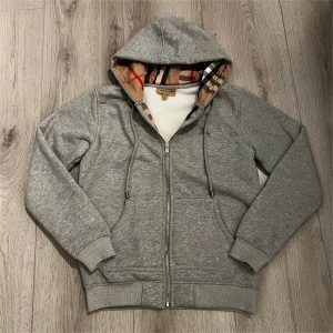 Grå Burberry hoodie med dragkedja - Snygg grå hoodie från Burberry med klassiskt rutigt foder i huvan och längs insidan. Tröjan har dragkedja framtill, snörning i huvan och ribbade muddar. Perfekt för dig som vill ha en stilren och lyxig look med ikoniska Burberry-detaljer.