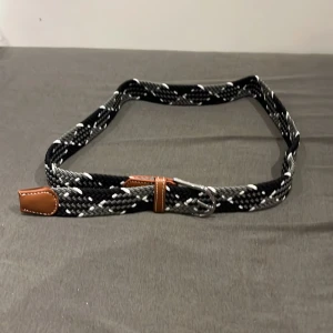 Svart och grått flätat bälte - Snyggt flätat bälte i svart, grått och vitt med bruna skinn-detaljer vid spännet och ändarna. Bältet har ett klassiskt silverfärgat spänne och är gjort i ett elastiskt material som ger en bekväm passform. Perfekt för att lyfta vilken outfit som helst. Helt ny och oanvänd